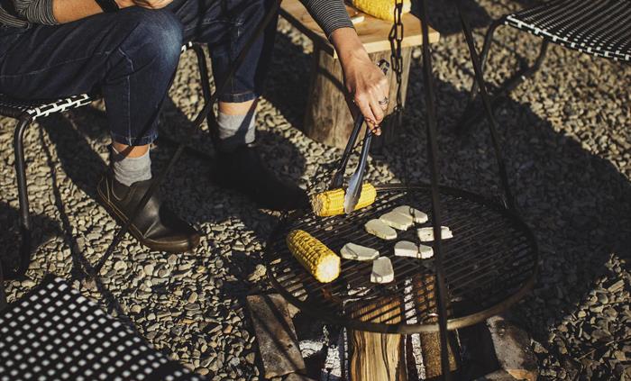 A close up of a campfire scene. A person uses tongs to turn corn on a barbecue hanging over a campfire.